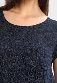 Námořnická modrá halenka vyrobená z měkkého materiálu, s kulatým výstřihem a bílým tečkovaným vzorem. Jsou viditelné krátké rukávy a hladký povrch.