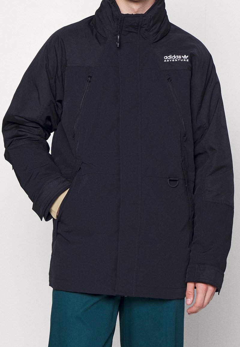 Svart, isolerad jacka med hög krage, dragkedjeförsedda bröstfickor och en logo på vänster sida. Har en lös passform och textil med struktur.