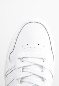 Bílé tenisky s hladkým koženým svrškem, plochými tkaničkami a elegantními stříbrnými akcenty po obvodu a na bočních pruzích.