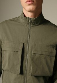 Mann trägt eine olivgrüne Jacke mit hohem Kragen, Frontreißverschluss und zwei großen Brusttaschen und steht vor einem einfarbigen Hintergrund.