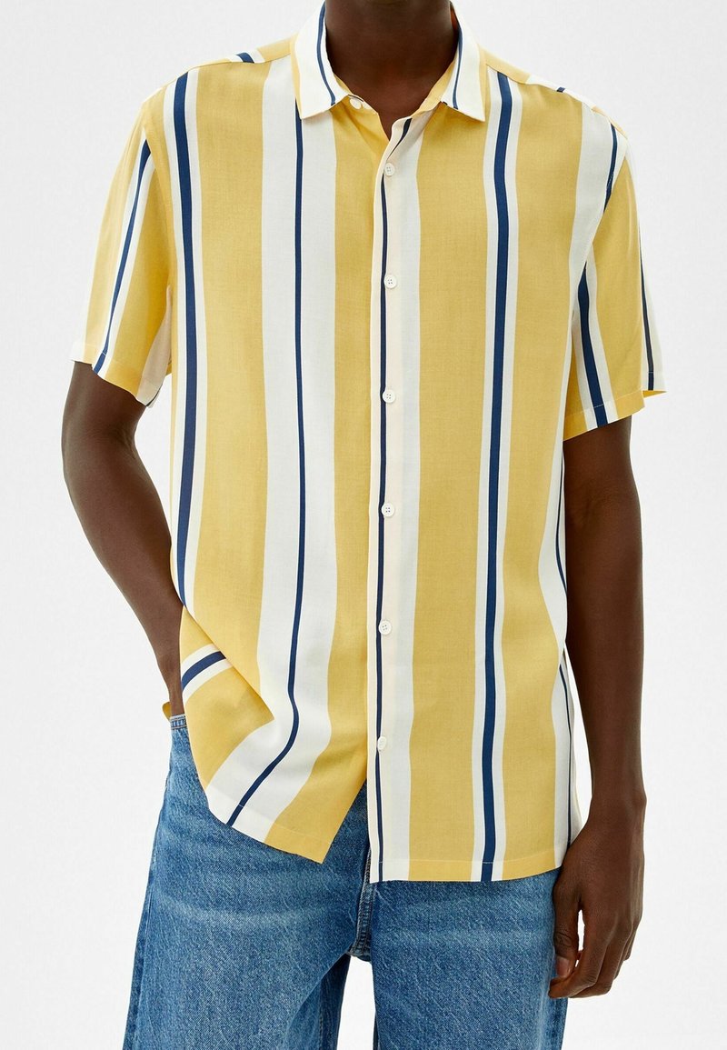 Hombre vestido con una camisa de botones de manga corta a rayas verticales en amarillo, blanco y azul marino, con jeans azules, una mano en el bolsillo, frente a un fondo blanco.