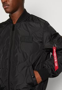 Mustan päällikankainen bomber-takki, jossa on vetoketjukiinnitys, ribbireunukset, logotarra ja punainen "Remove Before Flight" -tunnus hihassa.