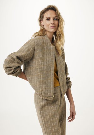 Veste bomber en cuir marron texturé avec une coupe décontractée, des fermetures à boutons et des poignets côtelés. Portée sur un haut moutarde et un pantalon à motif assorti.