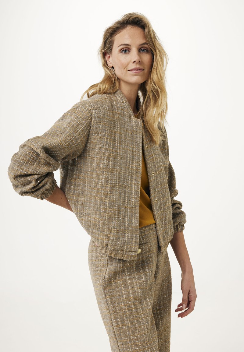 Brązowa, strukturalna kurtka bombowata w luźnym kroju, z zapięciami na guziki i ściągaczami przy rękawach. Noszona na musztardową górę i dopasowane spodnie w wzory.