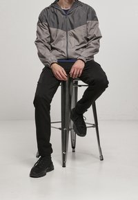 Person som sitter på en metalkrakk iförd en tvåfärgad grå jacka, svarta byxor och svarta sneakers mot en enfärgad ljus bakgrund.