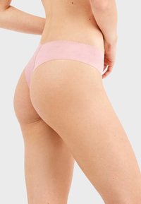 Tanga de algodón rosa claro con una textura suave y lisa y un diseño de cinturilla con ondas, vista desde un ángulo lateral.