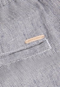 Gros plan d'un tissu texturé gris clair avec une petite étiquette beige portant la mention Jack & Jones au-dessus d'une couture soigneusement réalisée.