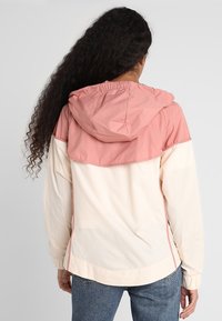 Kvinna med långt lockigt hår, iklädd en tvåfärgad rosa och krämfärgad jacka med huva samt blå jeans, vänd bortifrån mot en enfärgad bakgrund.