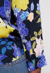 Květinová košile na knoflíky s námořnickým pozadím, živými modrými, žlutými a růžovými květinami, s hladkou texturou a detailem knoflíku na manžetě.