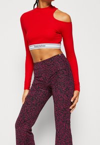 Punainen pitkähihainen crop-top, jossa on olkataskut ja valkoinen brändinauha. Mustat housut, joissa on vaaleanpunainen abstrakti kuvio ja levenevät lahkeet.