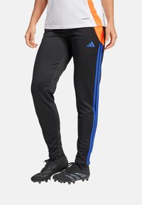 Schwarze Sporthose mit blauen Seitenstreifen. Hergestellt aus einem dehnbaren, glatten Material, mit passendem Design und schmalem Bein. Schwarze Fußballschuhe getragen.