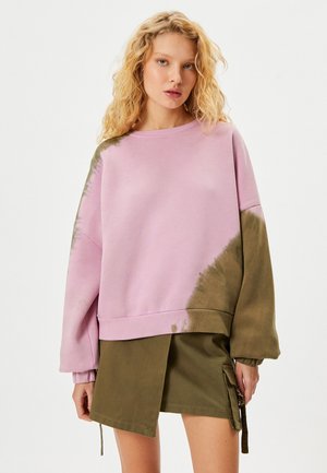 Yli suuri collegepaita vaaleanpunaisella ja vihreällä tahrakuosisella designilla. Puuvillamateriaali, ribbauskaihtimet ja helma, yhdistetty vihreään hameeseen, jossa on sivutasku.