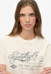 Ung kvinde med langt brunt hår iført en kridhvid Superdry t-shirt med vintage-inspireret logo og tekst.