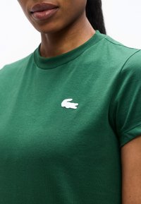 Γυναίκα που φοράει πράσινο t-shirt με ένα μικρό λευκό λογότυπο κροκόδειλου στο αριστερό στήθος, εμφανή τα χείλη και το πηγούνι, ουδέτερο φόντο.