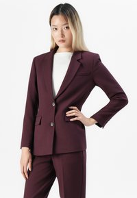 Kvinne med rett blondt hår som har på seg en tettsittende mørk lilla blazer og matchende bukser, poserer med en hånd på hoften mot hvit bakgrunn.