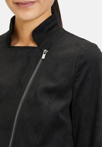 Zwarte jas met een hoge kraag, zilveren rits en een zachte textuur. Het ontwerp heeft een getailleerde vorm met verlengende naden.