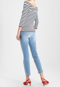 Bluzka odsłonięta na ramionach w biało-niebieskie paski, zestawiona z jasnoniebieskimi dżinsami. Modelka nosi czerwone szpilki. Widok z tyłu, minimalne tło.