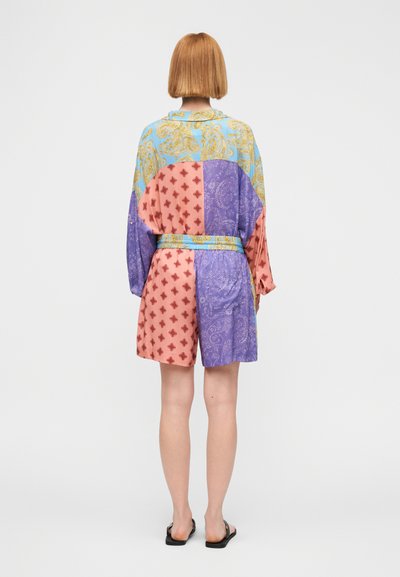 Osoba stojaca chrbtom k nám, oblečená v farebnej vzorovanej košeli a zodpovedajúcich kraťasoch, s čiernymi šľapkami na bielom pozadí.