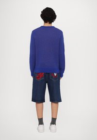 Suéter azul de malha com decote redondo, combinado com calções de ganga escuros com detalhes em vermelho nos bolsos traseiros. Ténis brancos.