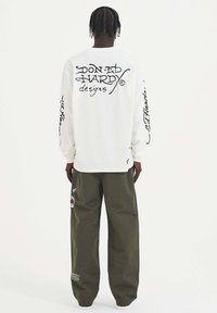 Witte lange mouwen shirt met zwarte cursieve tekst op de achterkant en mouwen, gecombineerd met olijfgroene wijde broek met patches en borduursels.