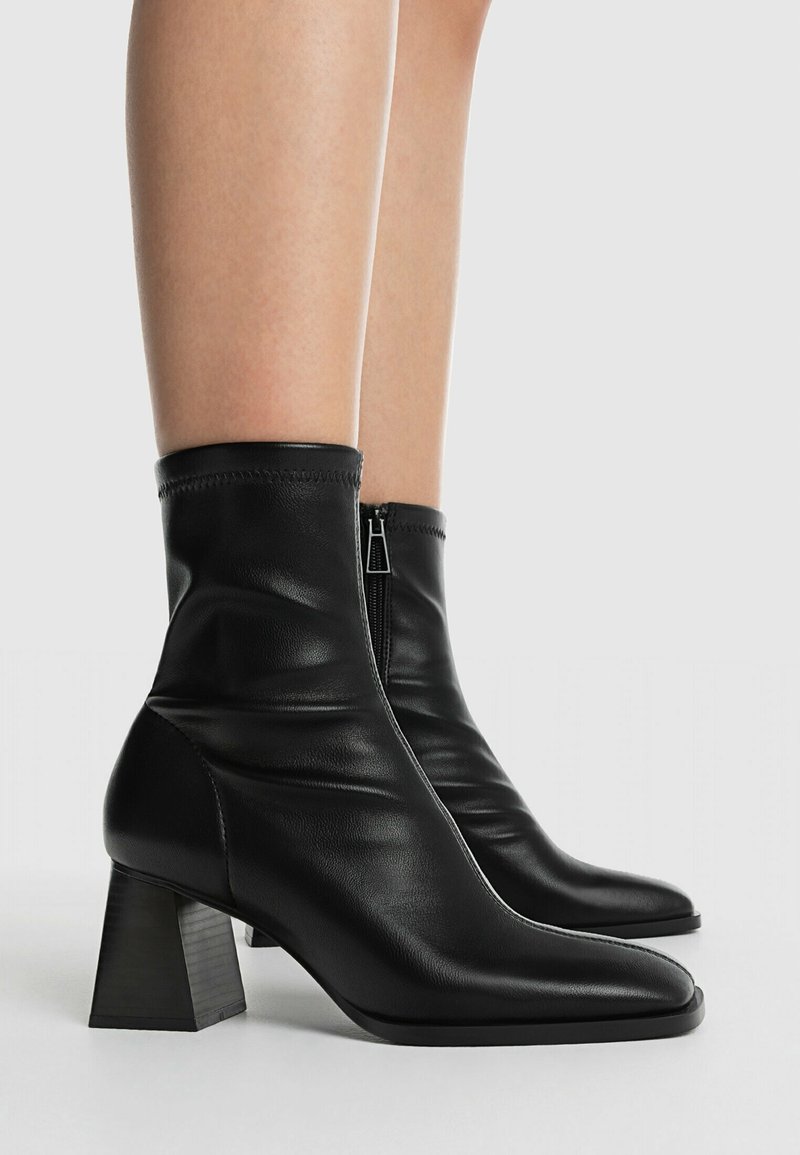 Bottes en cuir noir mi-mollet avec talons carrés et fermetures éclair latérales, portées sur des jambes nues, sur fond clair uni.