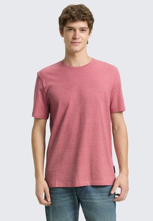 Jonge man met krullend haar, gekleed in een effen rood T-shirt met korte mouwen en een blauwe spijkerbroek, staand tegen een lichtgrijze achtergrond.