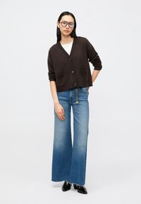 Rjav kratek kardigan z žepi in gumbi, kombiniran s širokimi modrimi jeans hlačami. Model nosi črne čevlje in očala; pod njim preprosta bela majica.