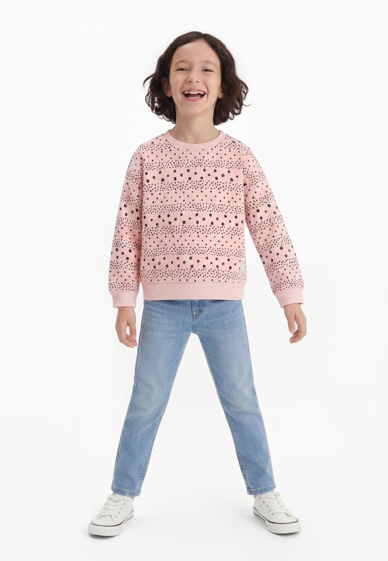 Smilende barn står med benene spredt iført en pink sweatshirt med prikker, lyseblå jeans og hvide sneakers mod en hvid baggrund.