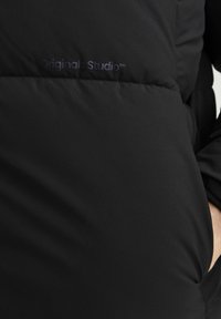 Musta polsterdatud jakk, millel on sile kangas, kergelt läikiv tekstuur ning tumesinise logo "Originals Studio" trükitud spordivormi ees.