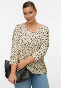 Chemise beige à col en V avec imprimé léopard noir, manches trois-quarts et coupe ample. Le modèle tient un sac à main noir contre un pantalon en denim.