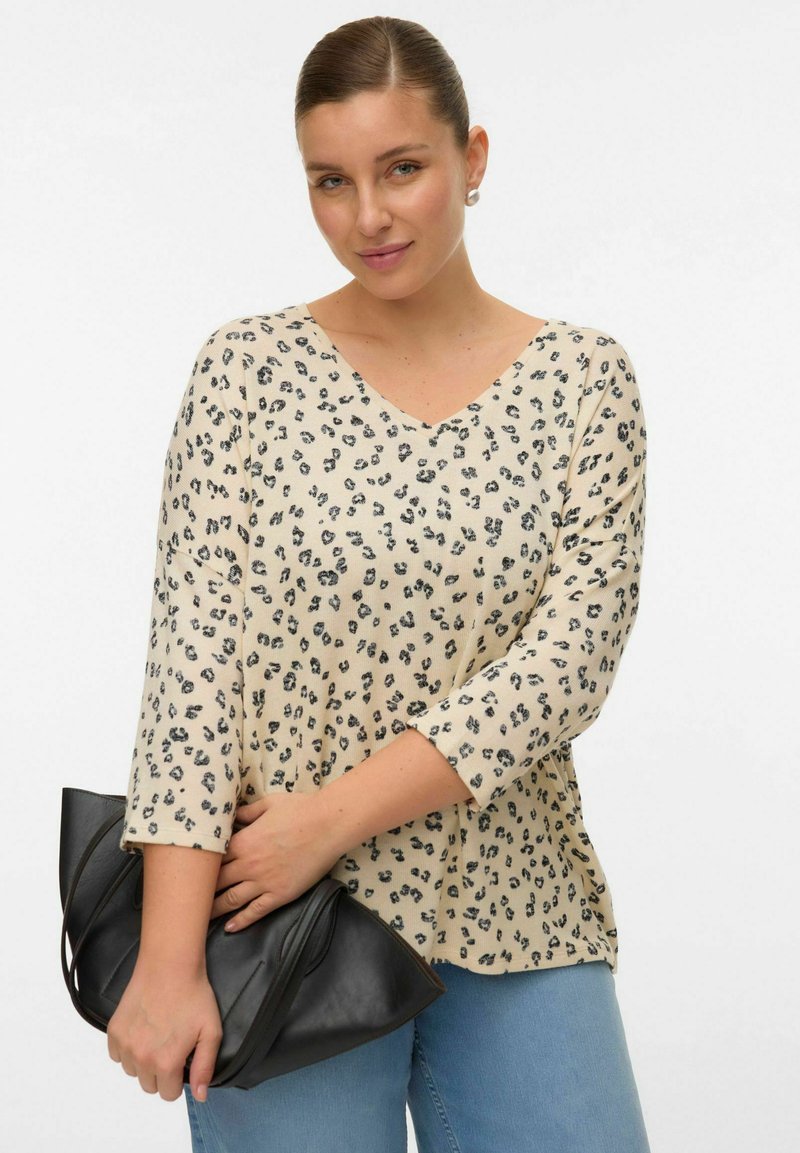 Chemise beige à col en V avec imprimé léopard noir, manches trois-quarts et coupe ample. Le modèle tient un sac à main noir contre un pantalon en denim.