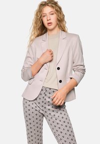 Helle graue Blazer mit schwarzen Knöpfen über einem beigen Oberteil, kombiniert mit gemusterten grauen Hosen, die ein sich wiederholendes Blumenmuster aufweisen.