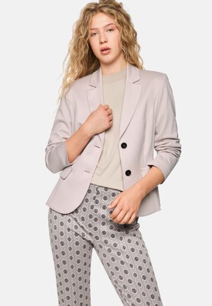 Lichtgrijze blazer met zwarte knopen over een beige top, gecombineerd met gestreepte grijze broek met een herhalend botanisch dessin.
