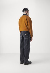 Brun langermet skjorte med svart krage, parret med mørk denim-bukser med to baklommer og et logo-patch. Svart sko.