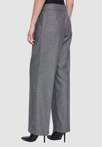 Donna che indossa pantaloni grigi a gamba larga a vita alta con tasche laterali e stivaletti neri con tacco, in piedi davanti a uno sfondo chiaro e semplice.