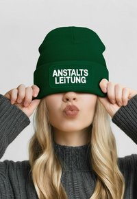 Green knit beanie with ribbed texture and a rectangular patch. The patch features the text "ANSTALTS LEITUNG" in bold white letters.