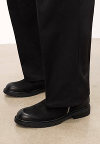Botas de cuero negras con punta redondeada, parte superior texturizada, suela de goma negra gruesa y un detalle de cremallera lateral. Llevadas con pantalones negros.
