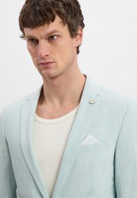 Hellblau strukturierter Blazer mit einer einzelnen Blumenbrosche, weißem Einstecktuch, getragen über einem cremefarbenen gerippten Hemd. Taillierte Passform, modernes Design.