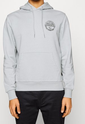 Homme portant un sweat à capuche gris clair avec poche avant et logo circulaire sur la poitrine gauche, associé à un pantalon noir sur fond uni.