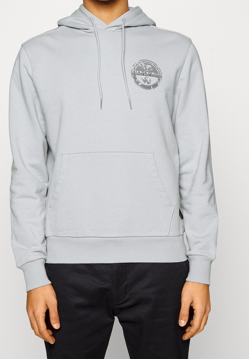 Homme portant un sweat à capuche gris clair avec poche avant et logo circulaire sur la poitrine gauche, associé à un pantalon noir sur fond uni.
