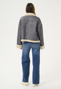 Grijze nep-shearlijk blazer met crème voering, oversized pasvorm, verlaagde schouders en contrasterende stiksels, gecombineerd met blauwe jeans.