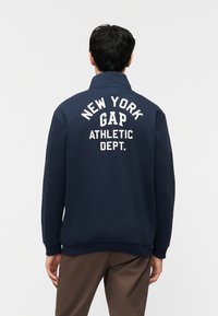 Άνδρας που φοράει μπλε φούτερ με την επιγραφή "NEW YORK GAP ATHLETIC DEPT." στην πλάτη, συνδυασμένο με καφέ παντελόνια, στέκεται με την πλάτη προς τα εμπρός.