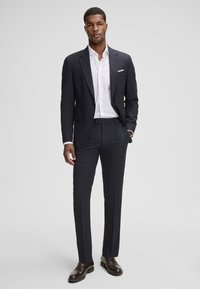 Marineblauer Anzug mit schlankem Schnitt, bestehend aus einem Sakko mit Revers und passenden Hosen, kombiniert mit einem weißen Hemd und dunkelbraunen Lederschuhen.