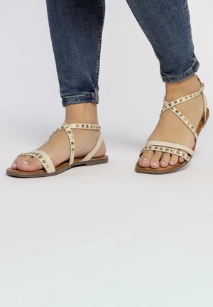 Beige platte sandalen met studs, voorzien van dunne banden en een minimalistisch ontwerp. Draagt met blauwe jeans, met open tenen.