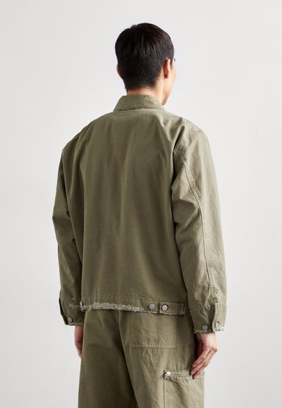 Olive green jacket with long sleeves, soft cotton material, frayed hem, and snap buttons at the cuffs and back, styled for a relaxed fit.