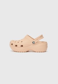 Light peach clogs made of synthetic material with ventilation holes, back strap, thick sole, and a decorative round logo accent.