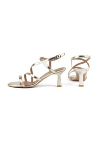 Goldene metallic High-Heelsandaletten besitzen kreuzende Riemen und eine quadratische Zehenpartie. Der Absatz ist ein metallischer Block mit einer strukturierten Oberfläche.