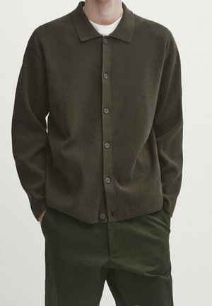 Homme portant un cardigan boutonné vert olive foncé sur un sous-vêtement blanc avec un pantalon vert olive foncé assorti, mains dans les poches, sur fond blanc uni.