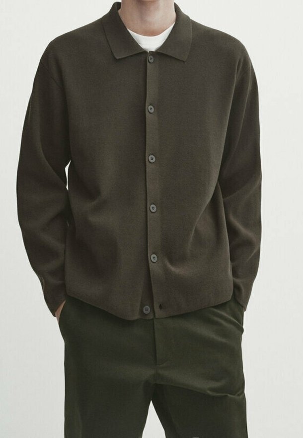 Homme portant un cardigan boutonné vert olive foncé sur un sous-vêtement blanc avec un pantalon vert olive foncé assorti, mains dans les poches, sur fond blanc uni.