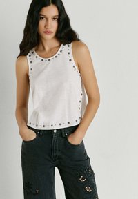 Ung kvinde med langt mørkt hår iført en ærmeløs hvid top med metalnitter og sorte jeans med blomsterudskæringsdesign.